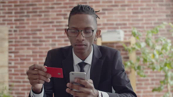 Black Man Office Worker in Glasses and an Official Suit Holds a Bank Card in Hand and Enters the