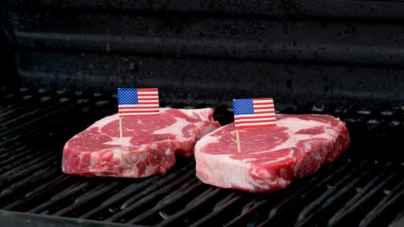 Two juicy rib-eye steaks sitting on the grill and cooking with two tiny American flags tooth picked