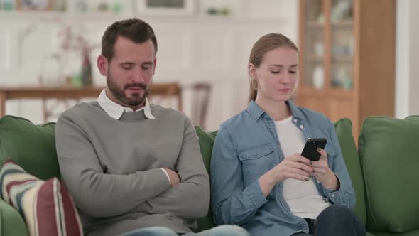 Woman Busy with Smartphone Sitting Next to Angry Man on Sofa