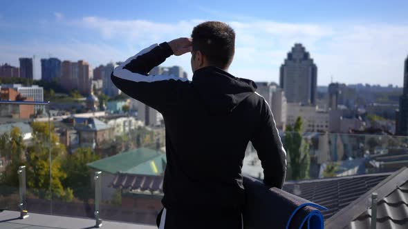 Sportsman with Exercise Mat Looking Around Holding Hand at Forehead Standing on Rooftop in Sunbeam