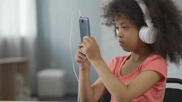 Serious African-American Kid Wearing Headset and Watching Video on Cellphone