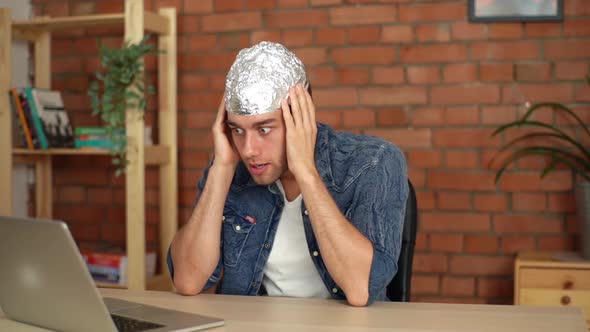 Closeup of Scared Conspiracy Theorist Wearing Foil Hat Watching Online Video Using Laptop Sitting at
