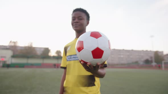 A Smiling Young Black Girl Stands in a City Stadium in Her Soccer Team Uniform and Holds the Ball