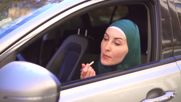 Muslim Woman in a Hijab Sits in a Car and Paints Her Lips with Lipstick