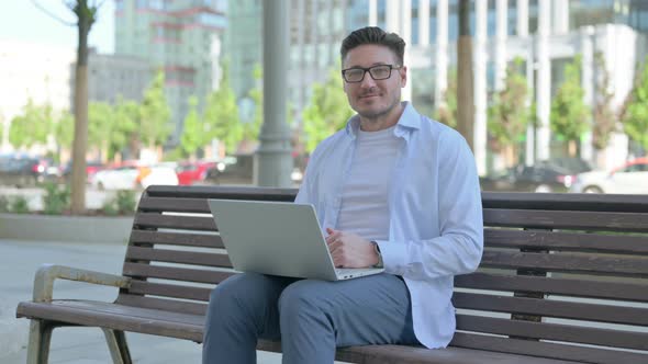 Man with Laptop Smiling at Camera While Sitting Outdoor on Bench