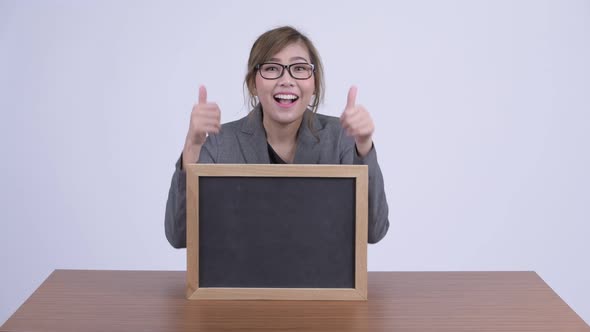 Young Happy Asian Businesswoman Showing Blackboard and Giving Thumbs Up at Desk