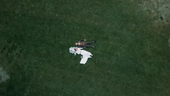 Beautiful young bride and groom lying on the grass holding hands in an amazing nature park