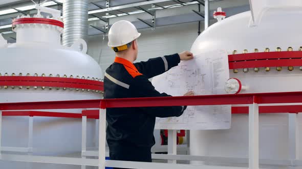 An Engineer in a White Helmet Holds a Drawing in His Hands Checks the Technological Equipment and