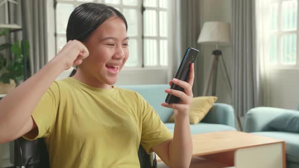 Happy Asian Kid Girl Sitting In A Wheelchair While Using Smart Phone And Celebrating At Home