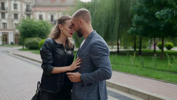 a Blonde in a Black Jacket and a Man with a Beard are Hugging Against a Background of Greenery