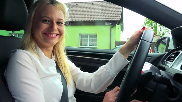 Young Attractive Blond Woman Drives a Car and Smiles To Camera While Waits on the Traffic Lights