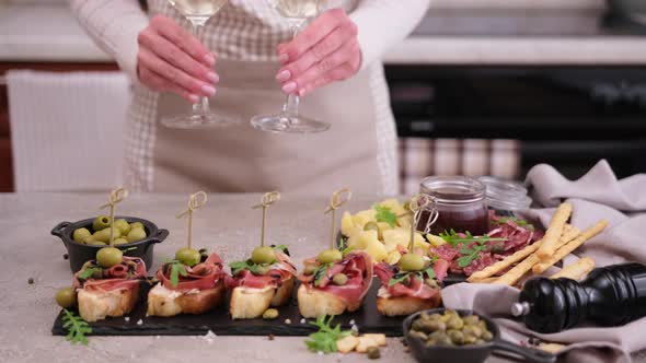 Woman Putting Two Glasses of Wine to Table with Meat and Cheese Plater at Domestic Kitchen