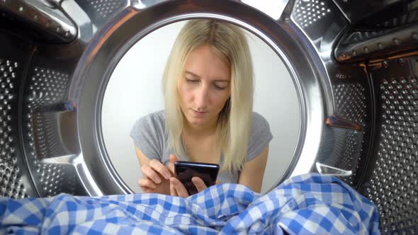 a woman looks at her smartphone through open door of washing machine.