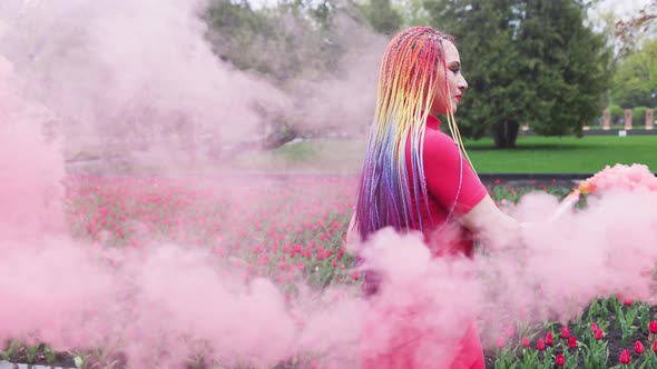 A Girl with Makeup with Rainbow Braids in Red Dress Posing in Red Smoke Against the Background of a