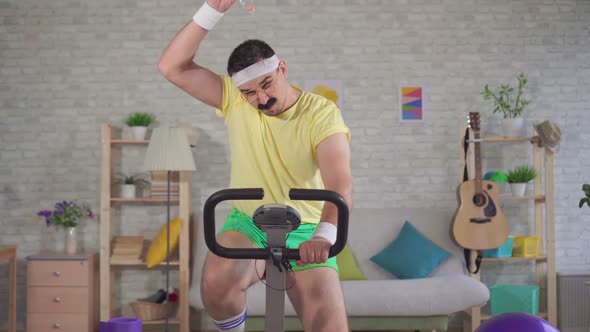 Funny Energetic Athlete of the 80's with a Mustache Is Engaged on a Exercise Bike and Pours Water