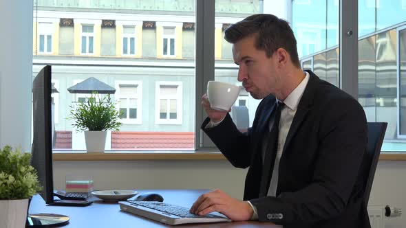 An office worker in a suit works on a computer and drinks coffee