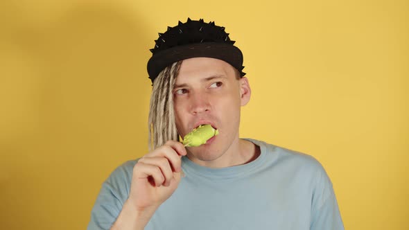 Young handsome man with dreadlocks in black cap eating ice cream on yellow background.