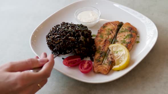 Dish with Fish and Black Rice on a White Plate. Woman Eating Fish Dinner in a Restaurant