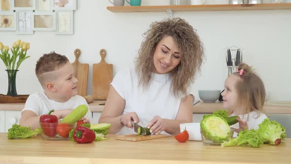 Young Blonde Woman with Curly Hair Cuts Vegetables and Communicates with Children Sitting at a Table