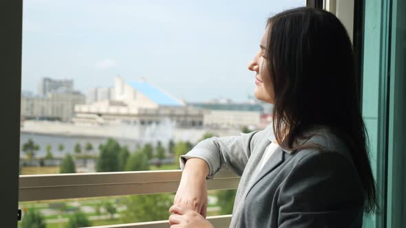 Young Woman in a Business Suit Stands By an Open Window and Looks Into the Distance