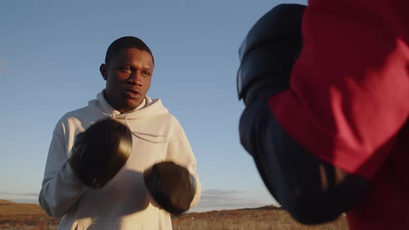 Black Man in Sportswear is Boxing with His Trainer Outdoors Front View