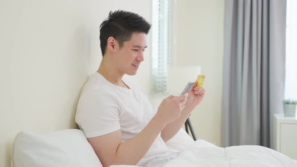 Asian attractive male enjoy shopping online on bed at home in morning.