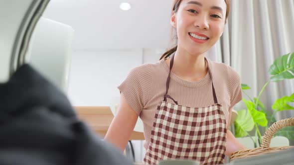 Asian young woman loads laundry in washer appliance, put dirty clothes to washing machine in house.