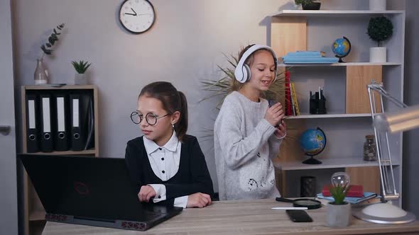 Teen Girl Dancing Under Inflammatory Music in Headphones and Another Younger Calm Girl Working