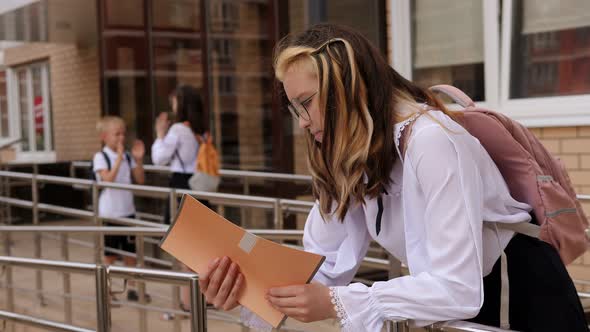 A Teenage Girl Reads Notes in a Notebook Standing in the School Yard