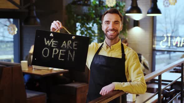 Happy Attractive Young Barista in Plaid Shirt and Brown Apron Holding Open Sign at the Coffee Shop