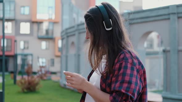 Attractive Girl Student in Headphones Looks at Smartphone Checking Social Networks Outdoors