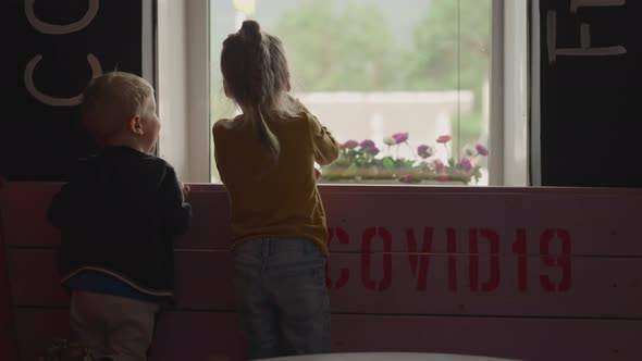 Little Girl Waves Hand and Has Fun with Brother Near Window