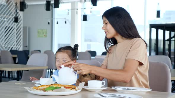 Mom Pours Tea From Teapot Into Cup Sitting By Girl and Pizza