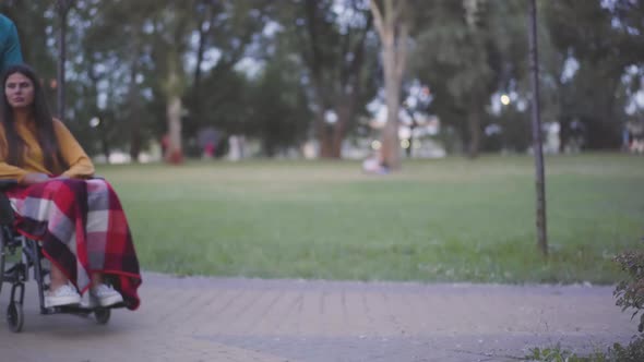 Side View of Man Rolling Wheelchair with Beautiful Disabled Woman in Park. Nurse or Volunteer and