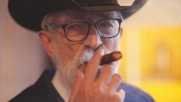 Stylish Old Man in Wide Brimmed Hat and Rich Blue Mens Suit Smoking Cigar Indoor