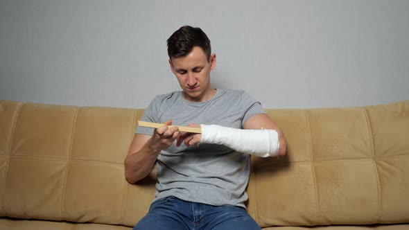 Young Man Scratches Forearm in Gypsum Band Frowning on Sofa