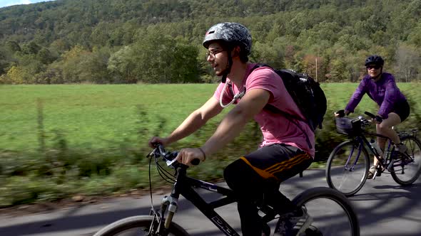 Teenage boy biking with helmet and glasses on a rural road with farmhouse and mother on bike in the