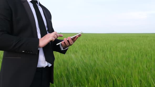 A Man in a Black Business Suit Uses a Tablet Among the Green Field