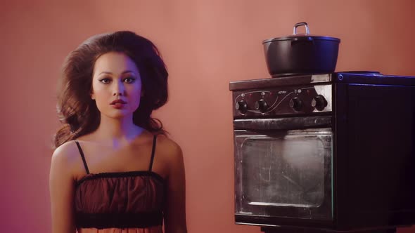 Beautiful Asian Girl Sits in Front of Oven on Bright Background and Waits for Preparation of Dish