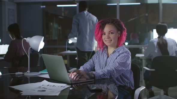 Creative Manager with Pink Hair Typing on Laptop and Smiling at Camera Working Late in Office