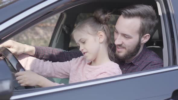 Little Girl Sitting on Father's Lap in the Car Holding Wheel Close Up. The Child Is Learning To
