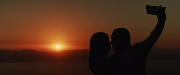 Silhouette of a young couple taking a selfie at sunset, while kissing