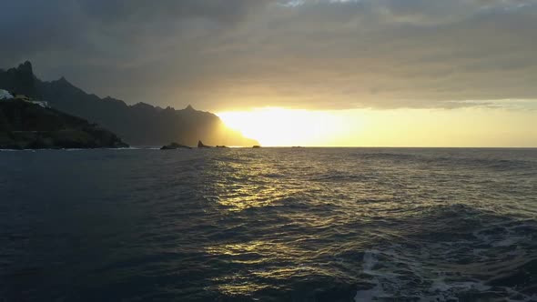 Backward Flight Above Ocean Waves Facing Beautiful Sunset and Mountains on Horizon. Aerial Drone