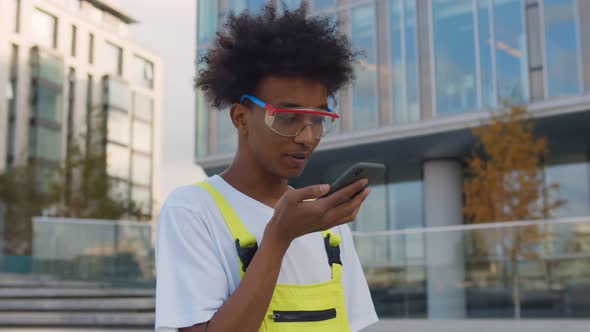 Young African Worker in Overall and Glasses Recording Voice Sms on Smartphone Standing Outdoors