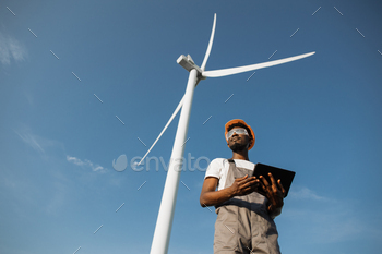 ntrolling working process of wind turbines outdoors. African american man wearing grey overalls, orange helmet and safety glasses.