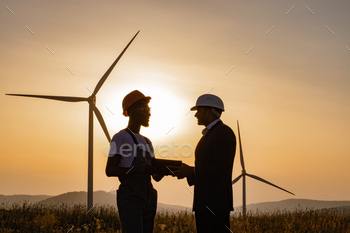  inspector using digital tablet during meeting outdoors. Two partners standing on field with wind turbines during amazing summer sunset.