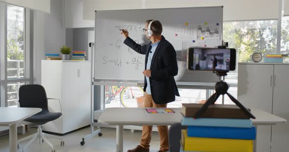 Mature Physics Teacher in Headphones Writing on Whiteboard Making Tutorial Video on Smartphone