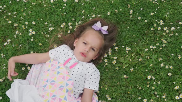 Little Happy Child Girl in Dress with Bow on Head Laying on Green Lawn in the Park