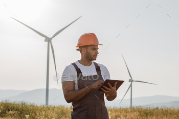 ith wind turbines and using digital tablet. Competent engineer wearing orange helmet and grey overalls.