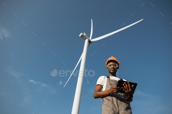 ntrolling working process of wind turbines outdoors. African american man wearing grey overalls, orange helmet and safety glasses.